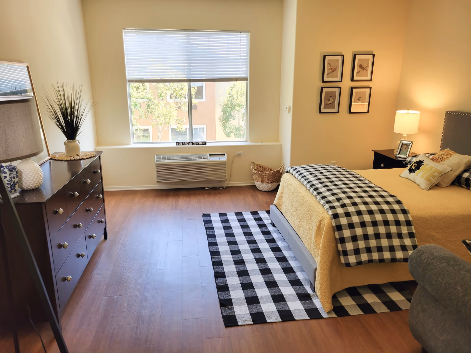 A bright bedroom with a large window letting in natural light. The room features a bed with a yellow bedspread and a black and white checkered blanket and rug. There is a dark wooden dresser with decorative items including a lamp and a plant. On the wall above the bed are four framed pictures of birds. A small basket is placed near the window, and a nightstand with a lamp and picture frame is beside the bed.