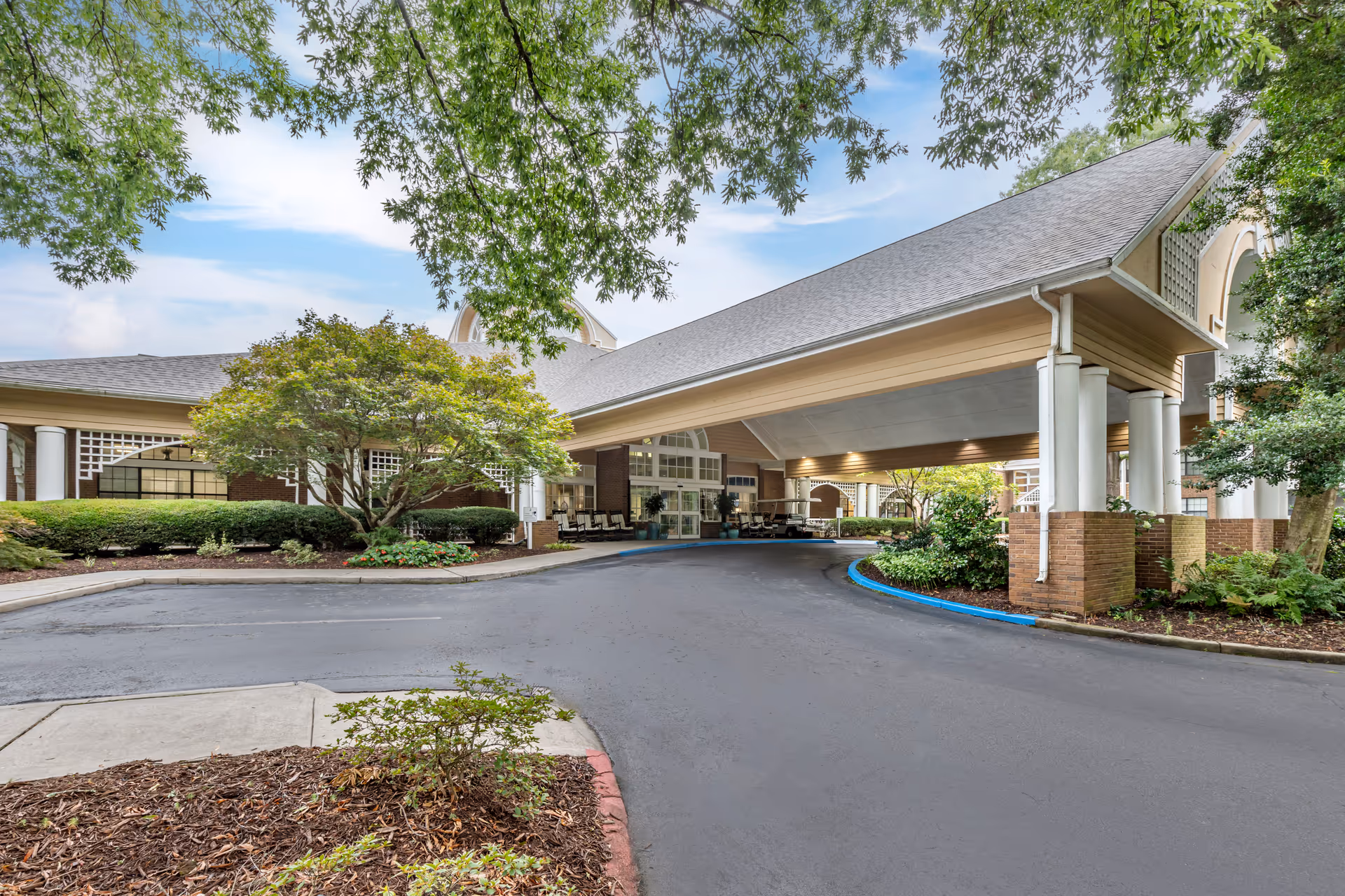 Entrance of a senior living facility with a covered driveway, surrounded by landscaped greenery including trees and bushes. The building features brick and white columns with a large roof extending over the driveway.