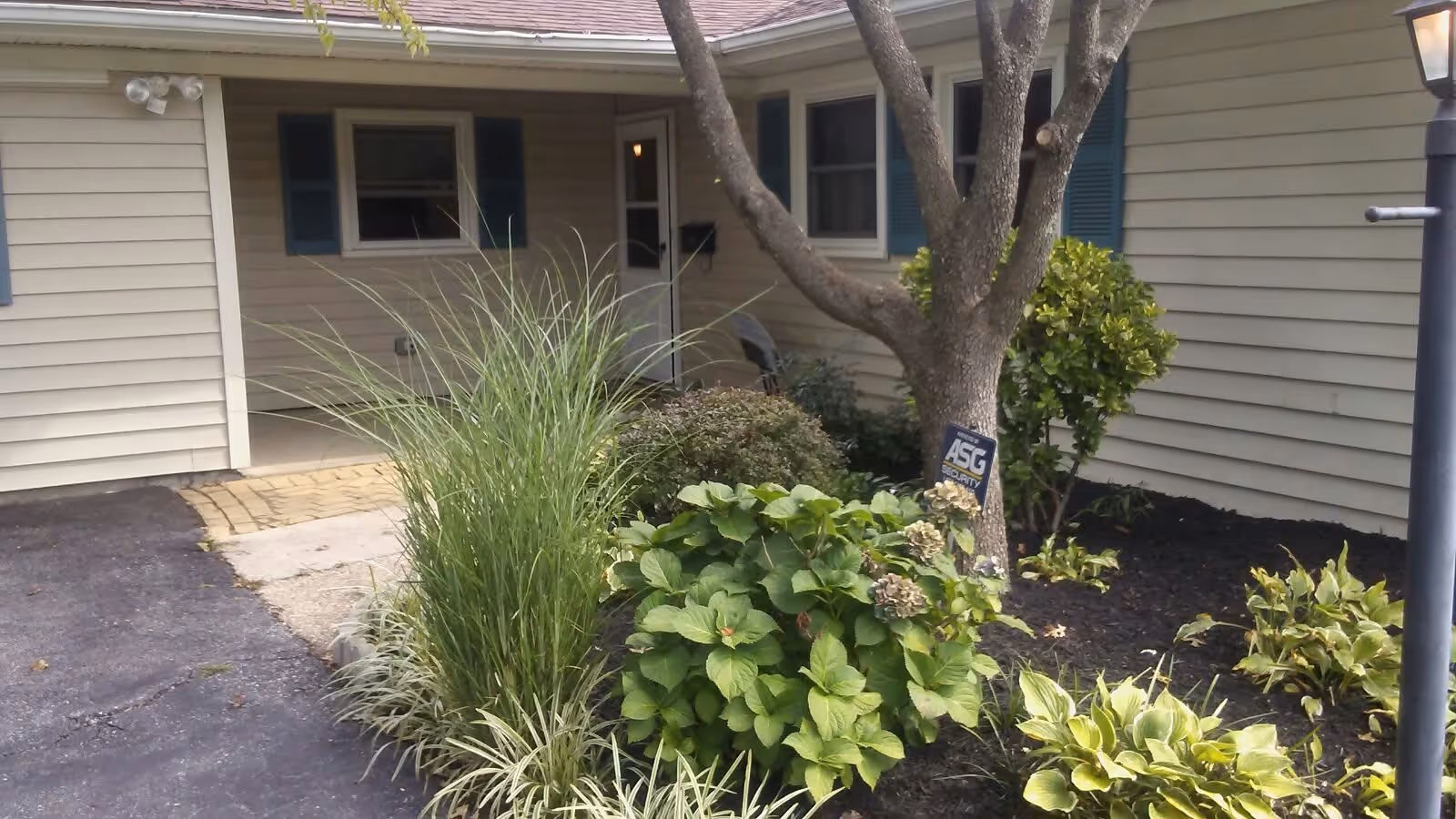 Exterior view of a single-story building with beige siding and blue shutters. There is a small garden area with various green plants and a tree in front of the building. A paved driveway or walkway is visible on the left side, and a lamp post is on the right side.