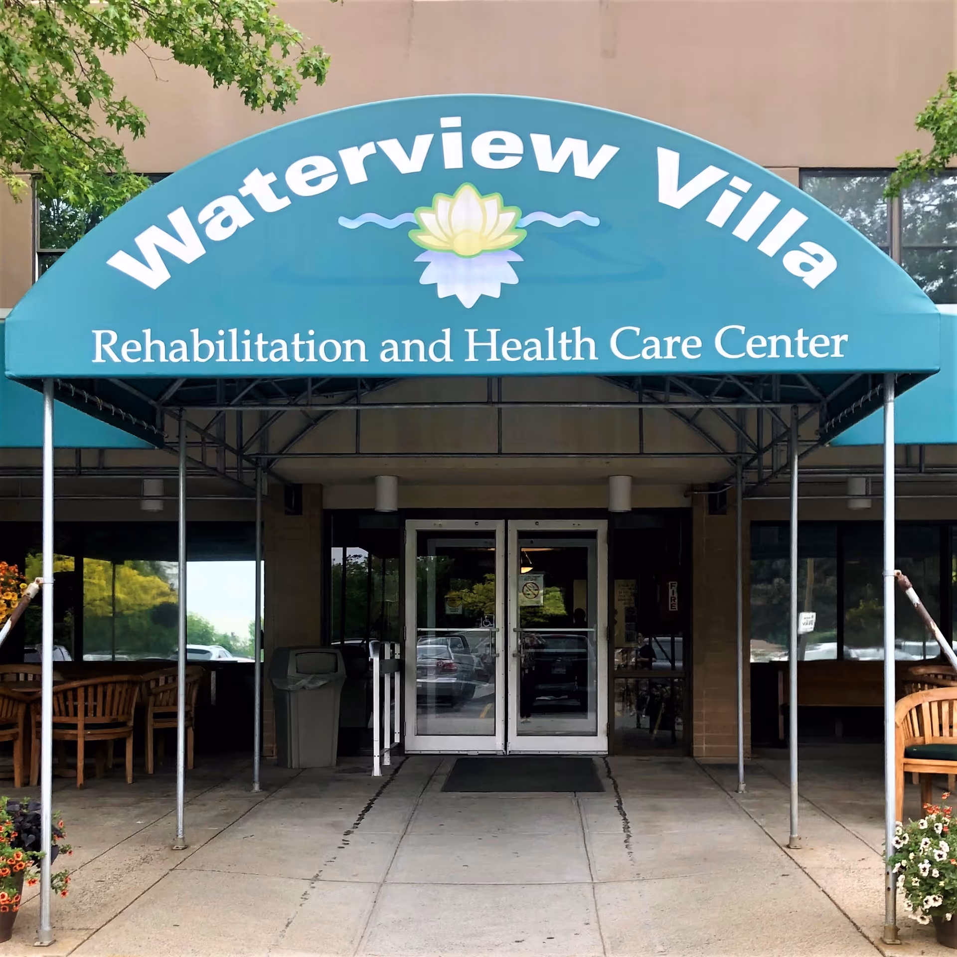 Entrance to Waterview Villa Rehabilitation and Health Care Center with a teal awning displaying the facility name and logo above double glass doors. There are benches and potted plants on either side of the entrance.