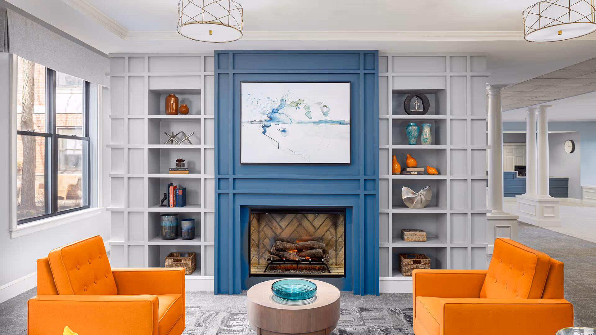 A cozy living room area featuring two bright orange armchairs facing a blue fireplace with a modern abstract painting above it. The fireplace is flanked by white built-in shelves decorated with various vases, books, and decorative objects. Large windows on the left allow natural light to fill the space, and the room has a light gray carpet with a subtle pattern. In the background, white columns and a reception desk area are visible.