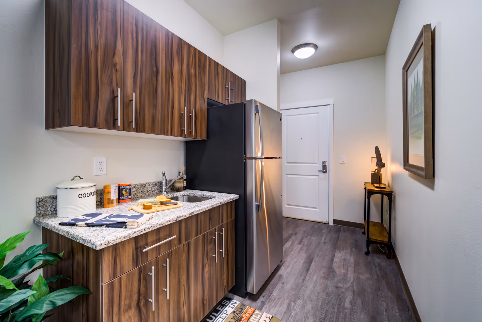 A small kitchen area with wood-grain cabinets, a granite countertop, a stainless steel refrigerator, and a sink. On the countertop, there is a cookie jar, some packaged food items, a cutting board with cheese and bread, and folded kitchen towels. The floor is wood-style laminate, and there is a small table with a lamp and a framed picture on the wall near a white door.