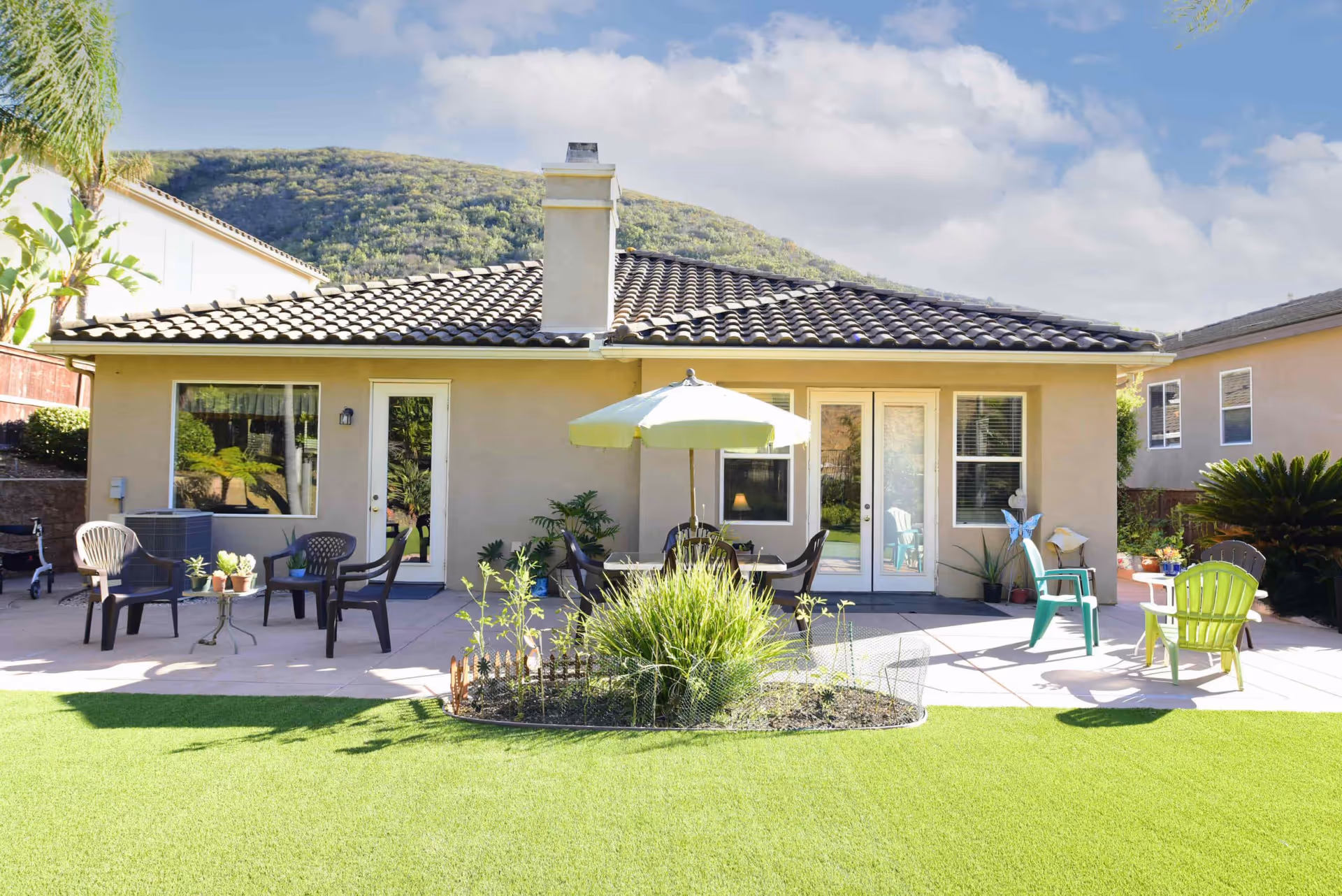 Sunny backyard patio of a single-story stucco building with a tiled roof, chairs, umbrella, potted plants and artificial turf with hills in the background.