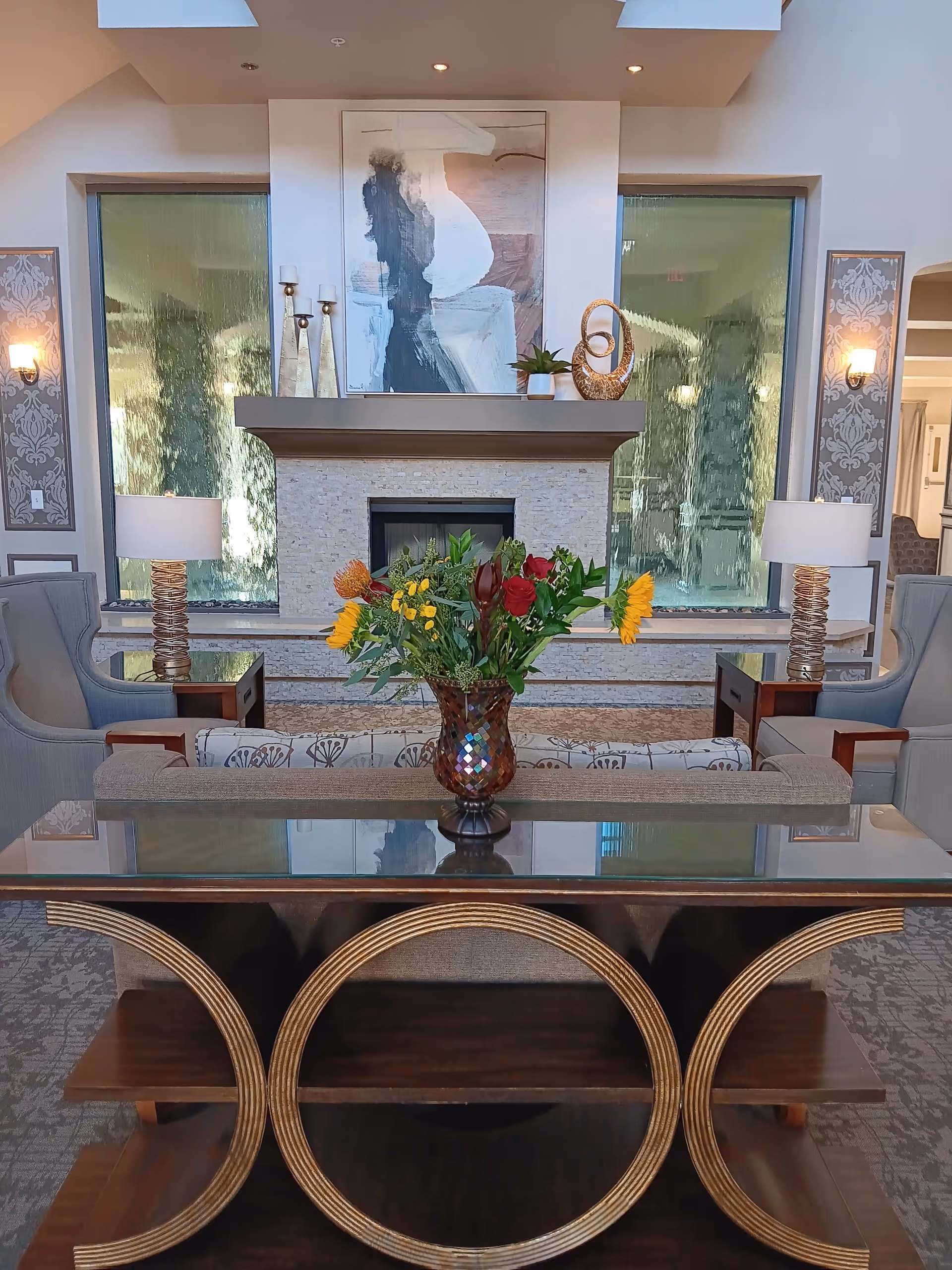 A senior living lounge with a glass-top console table and vase of flowers centered in front of a fireplace and seating area.