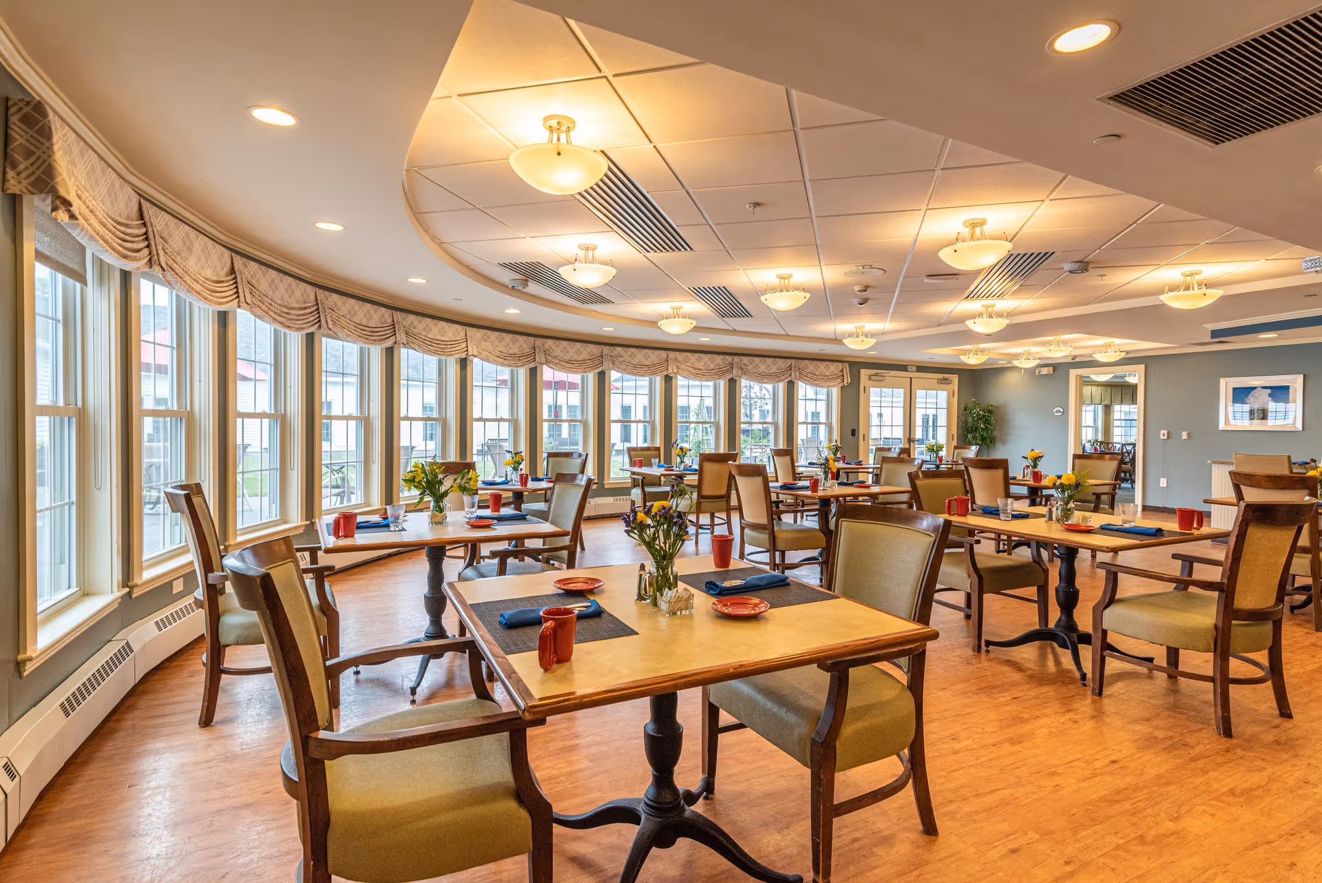 A bright and spacious dining room with multiple wooden tables and chairs arranged neatly. Each table is set with red mugs, blue napkins, and small flower vases. Large windows with beige valances line the curved wall, allowing natural light to fill the room. The ceiling has recessed lighting and decorative light fixtures, and the floor is a warm wood tone.
