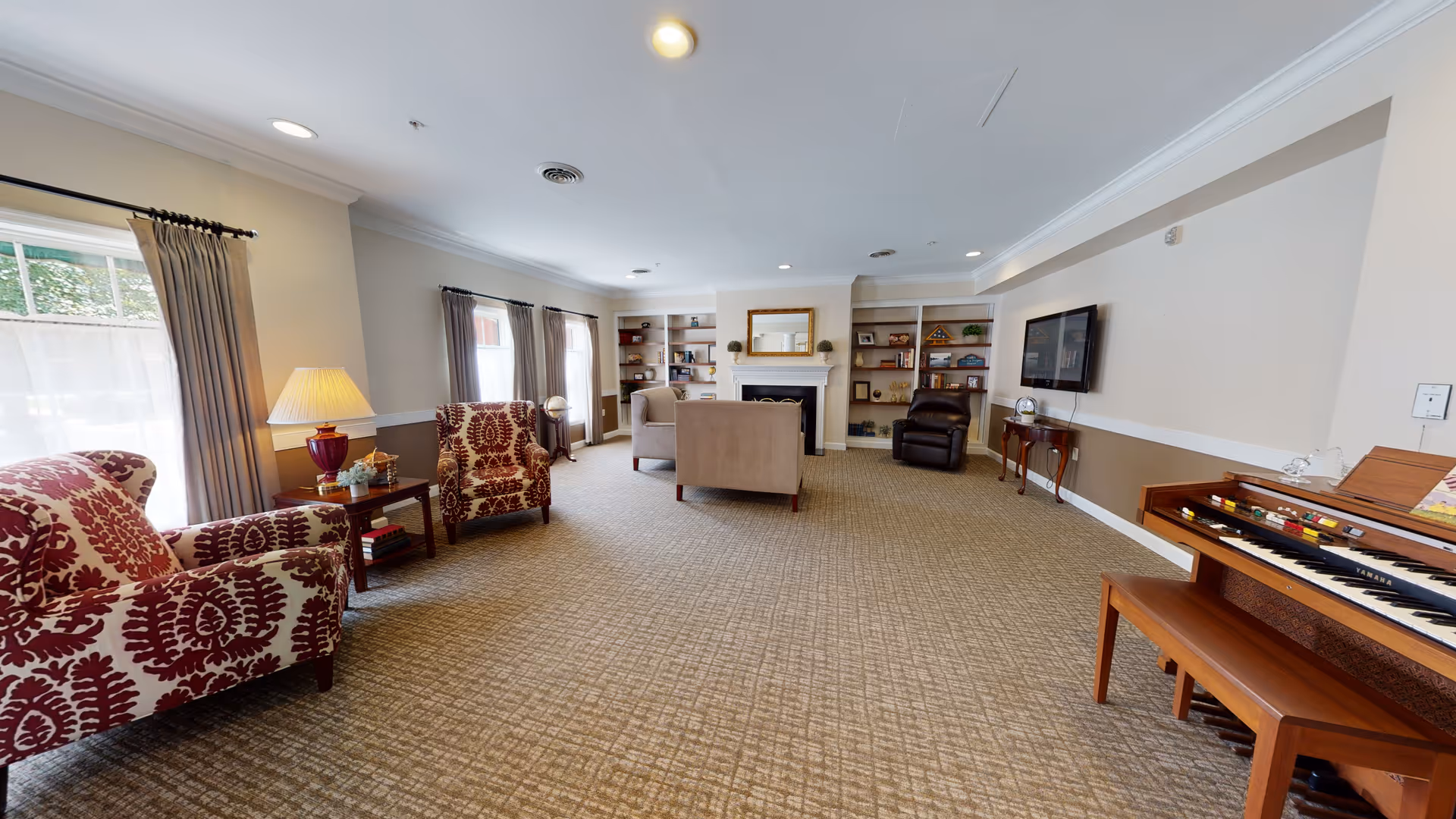 Bright communal living room with patterned armchairs, a sofa facing a fireplace and built-in bookshelves, a wall-mounted TV, and a piano.