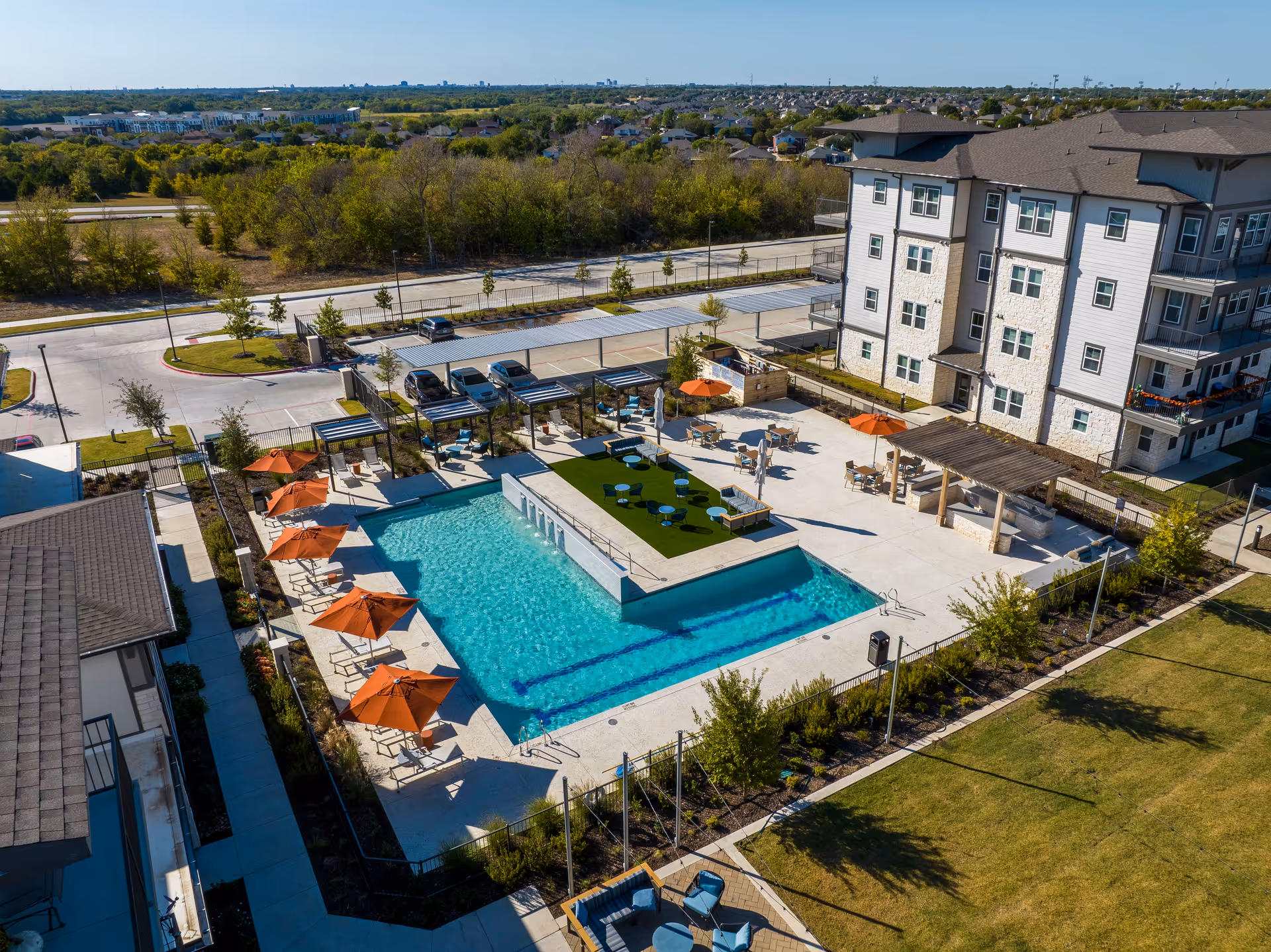 Aerial view of an outdoor pool area at Amberlin at The Station, featuring a rectangular swimming pool surrounded by lounge chairs with orange umbrellas, seating areas with tables and chairs, pergolas, and a multi-story residential building in the background. The area is fenced and landscaped with trees and shrubs, with a parking lot and greenery beyond.