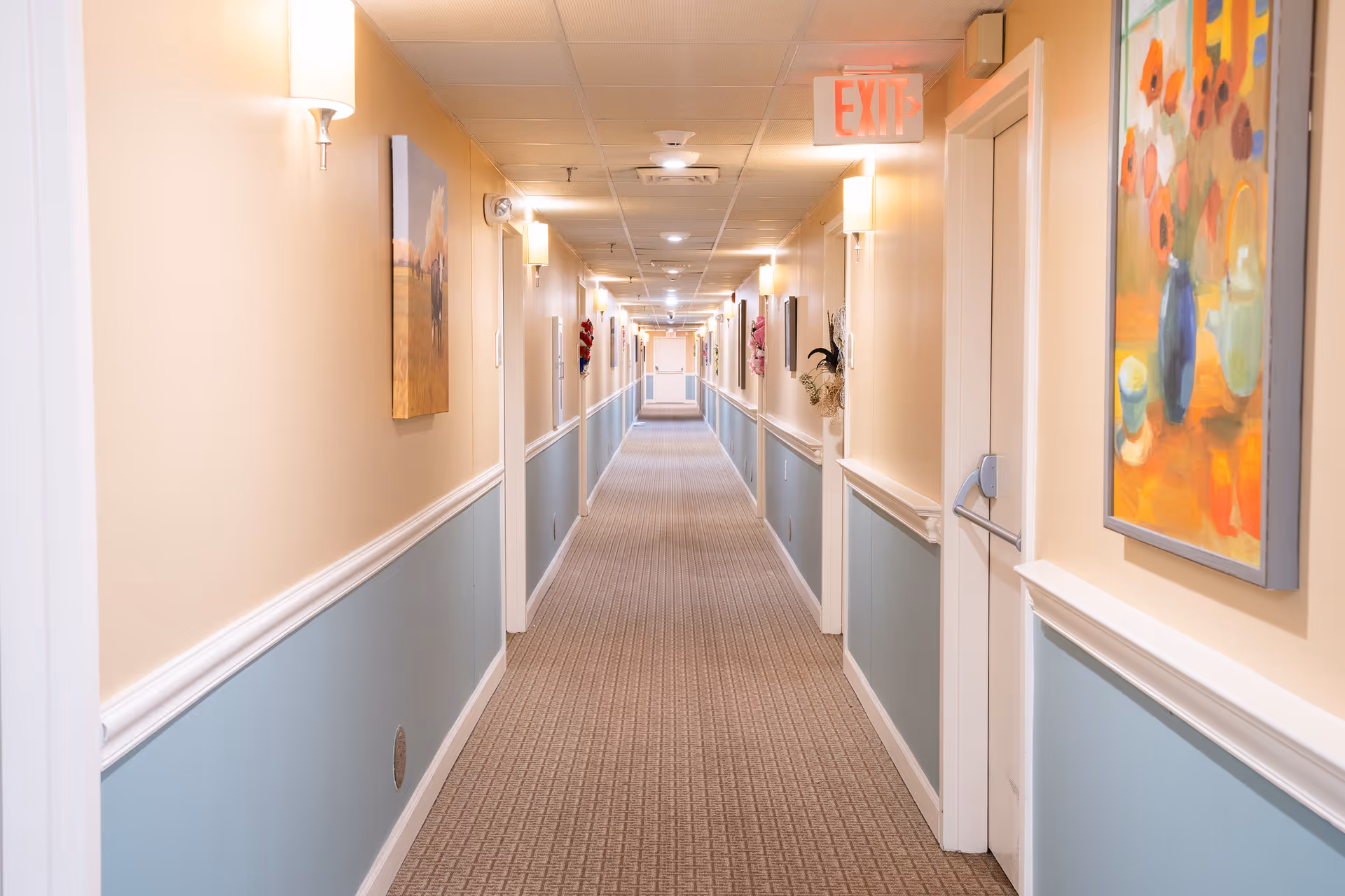 A long, well-lit hallway in a senior living facility with beige and light blue walls, carpeted floor, wall-mounted lights, and colorful paintings on the walls. Several doors line the hallway, and an illuminated exit sign is visible on the ceiling.