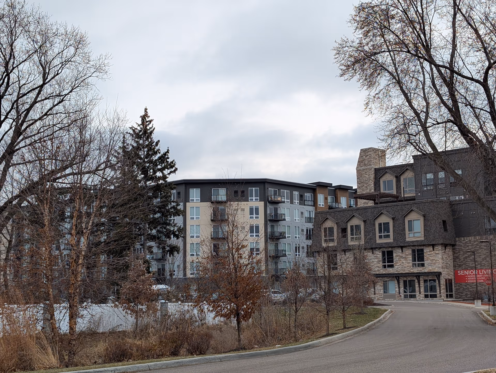 Exterior view of a senior living facility building with multiple floors, balconies, and a stone facade section. Leafless trees and a curved driveway are visible in the foreground under a cloudy sky.