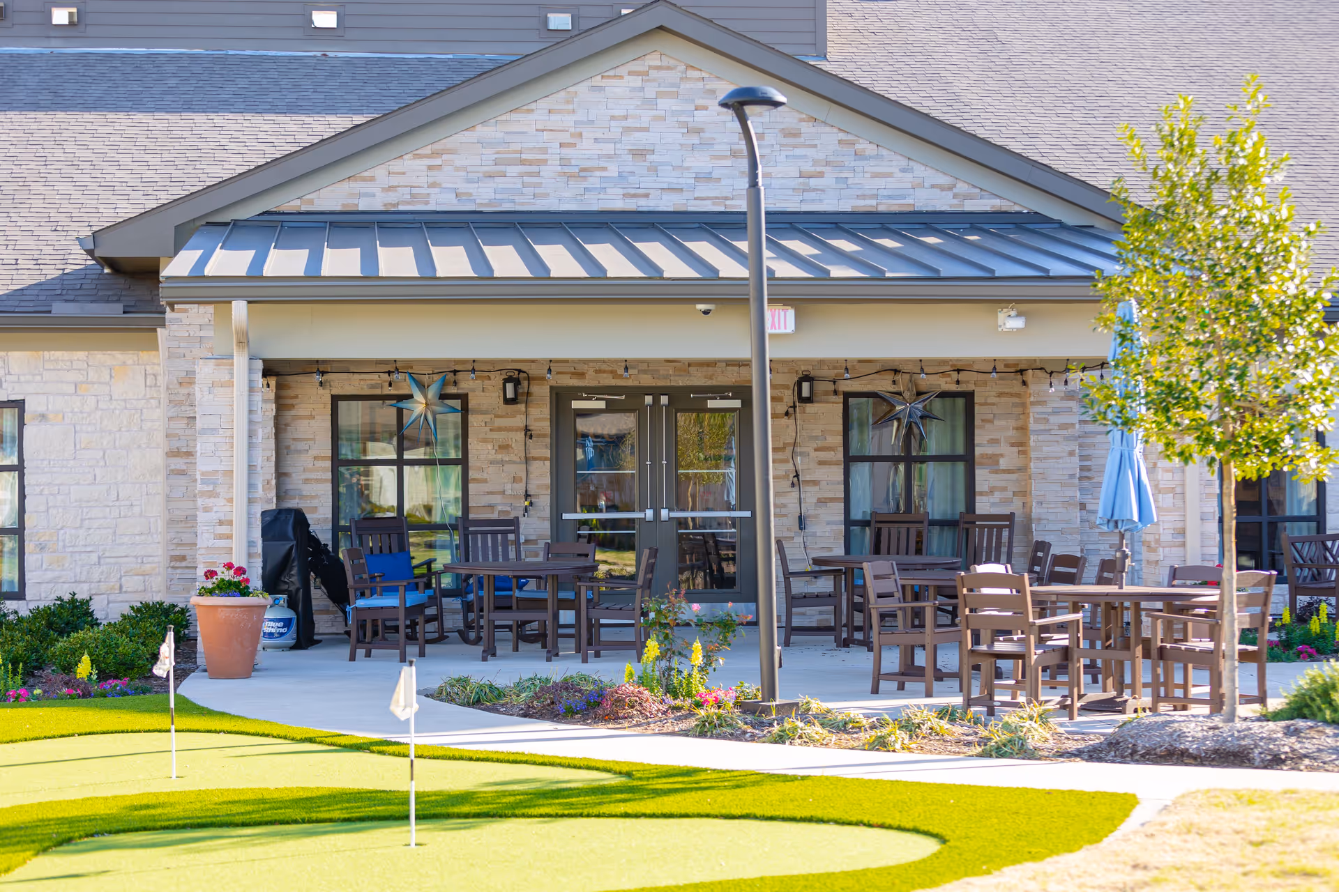 Outdoor patio area at The Village at Sugar Land with several wooden tables and chairs, a small tree, potted plants, and a putting green in the foreground. The building has stone walls and a metal roof over the patio.