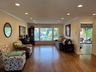 Common area living room with armchairs, a sofa, hardwood floors, a large front window and recessed ceiling lights.