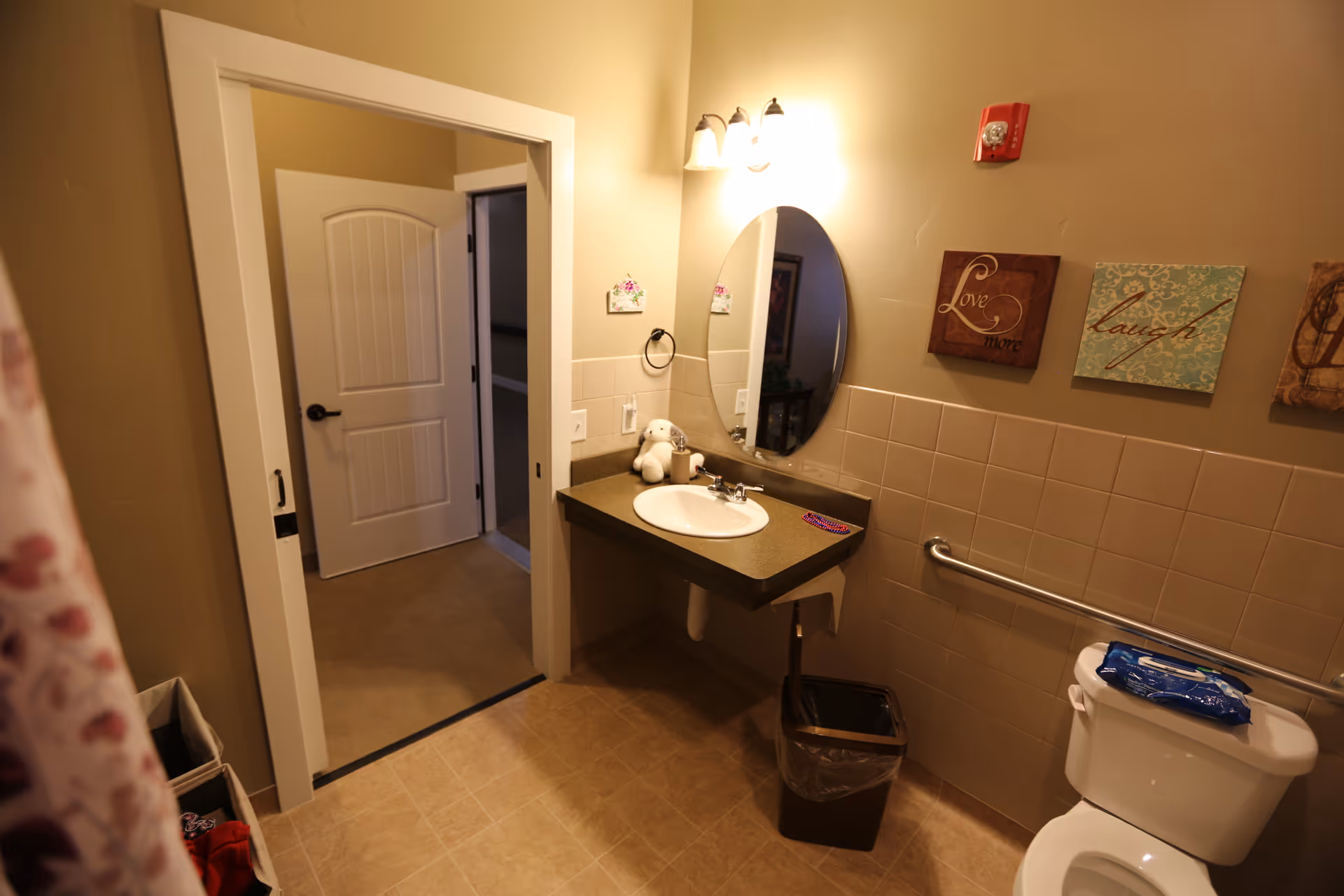 A bathroom with beige walls and tiled floor featuring a white toilet with a pack of wipes on the tank, a countertop with a sink, an oval mirror above the sink, a small stuffed animal, and wall decorations with the words 'Love' and 'Laugh'. There is a door leading to another room and a trash can under the sink.