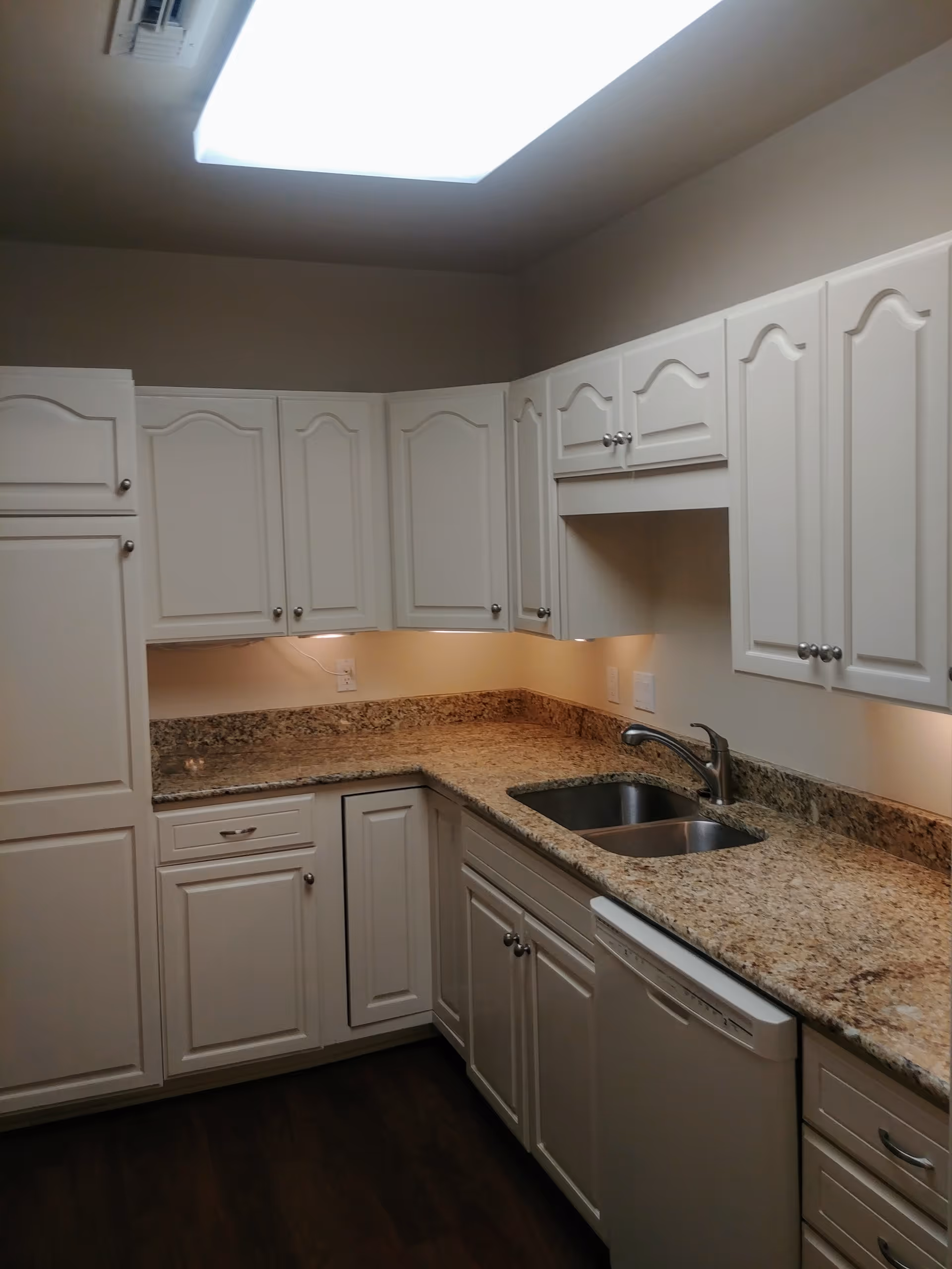 A kitchen with white cabinets, granite countertops, a double stainless steel sink, and a dishwasher. The kitchen has under-cabinet lighting and a large ceiling light fixture.