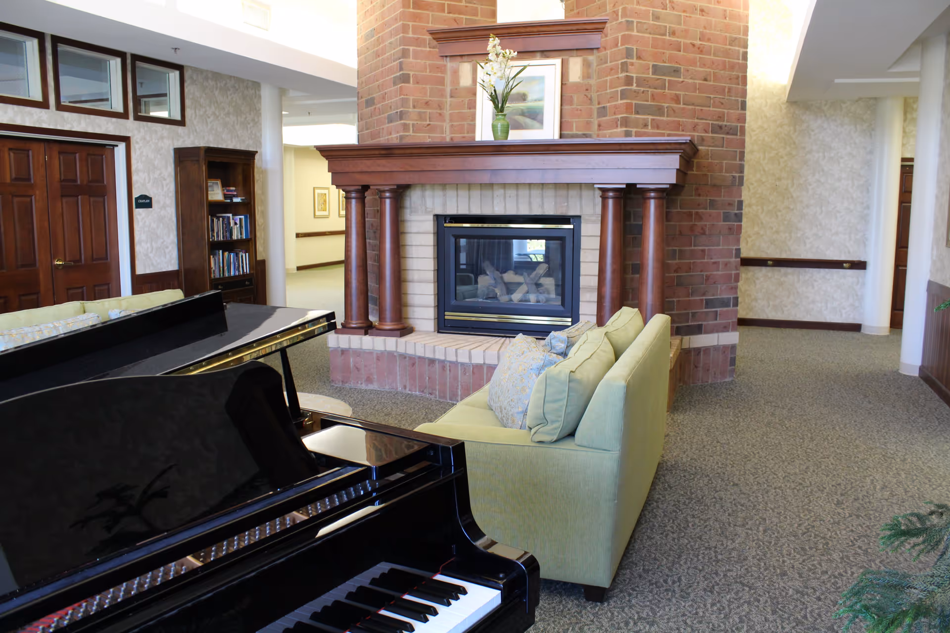 Interior of a senior living facility lounge area featuring a black grand piano in the foreground, a green sofa with decorative pillows, and a brick fireplace with wooden mantel and a vase with flowers on top. There is a bookshelf and wooden double doors in the background.