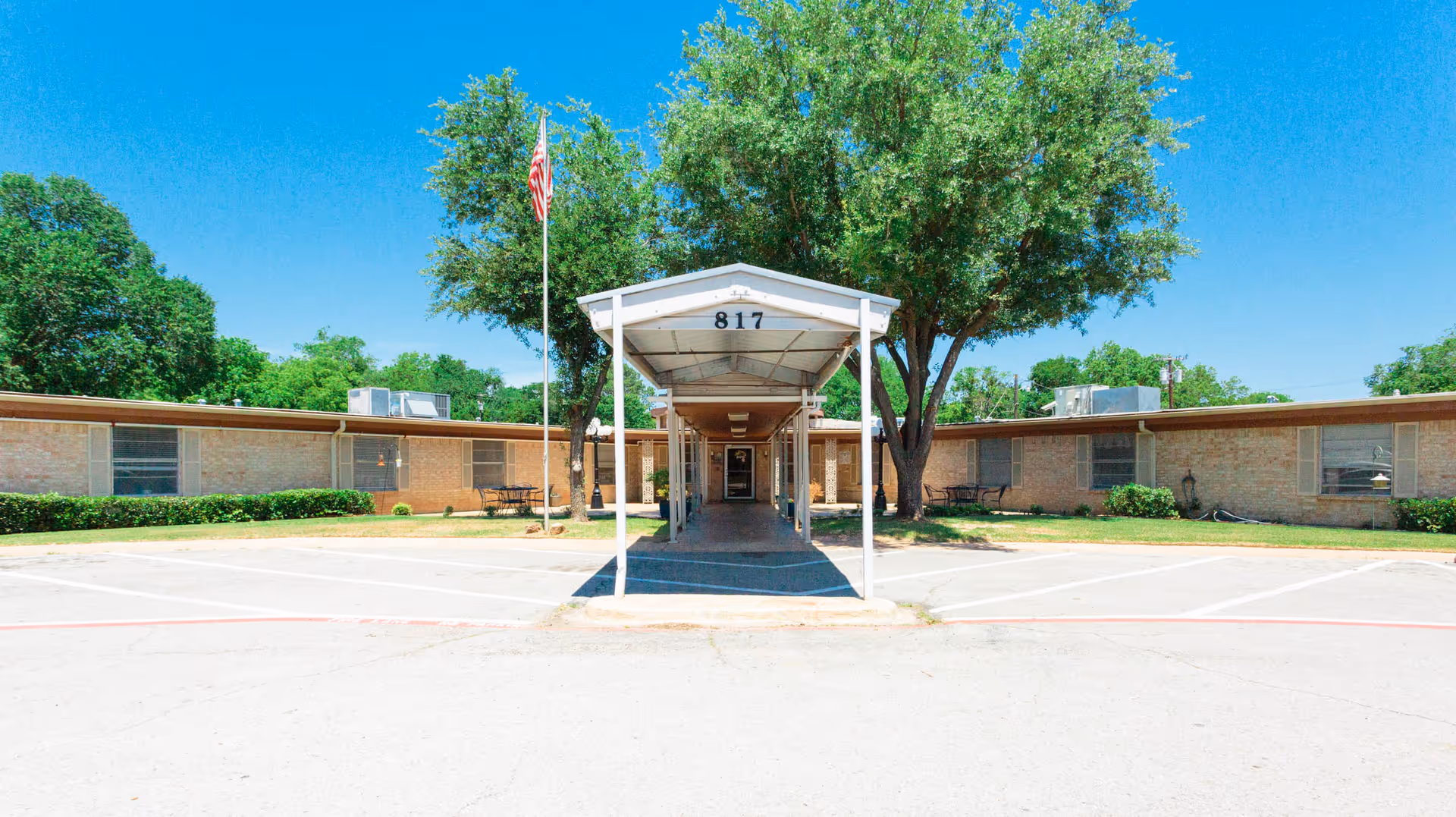 Front exterior view of a single-story brick building with a covered walkway leading to the entrance. The building has the number 817 displayed above the walkway. There are trees and bushes around the building, an American flag on a flagpole to the left of the walkway, and parking spaces in front.