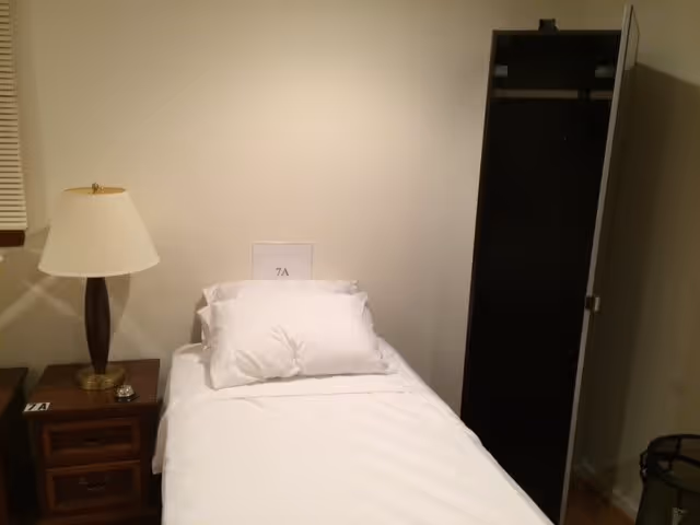 A small bedroom with a single bed made with white sheets and pillows. There is a wooden nightstand with a lamp on it to the left of the bed. A sign with '7A' is placed on the bed. To the right, there is an open closet with a mirrored door.
