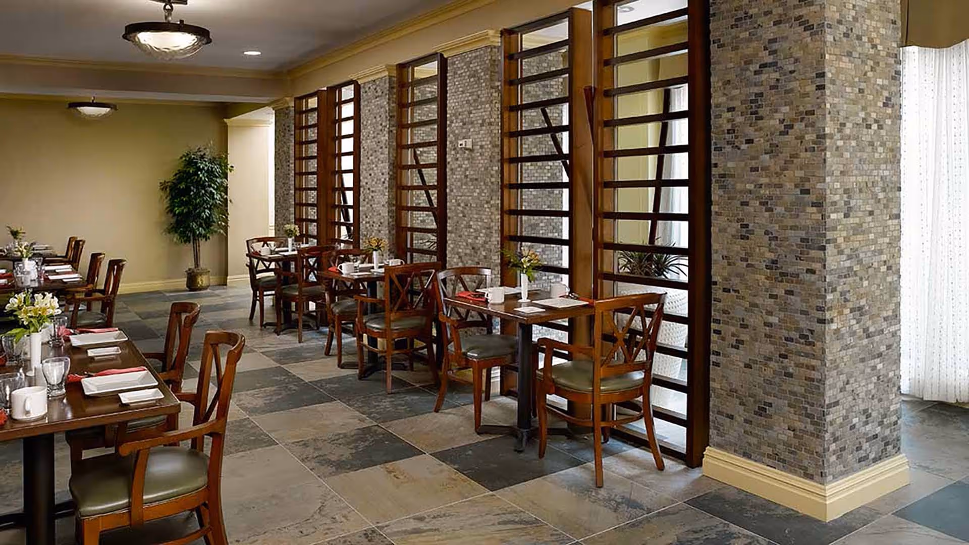 A dining room with several wooden tables and chairs arranged neatly. Each table is set with white plates, cups, glasses, and napkins. The room features a tiled floor, stone-textured columns, and wooden lattice dividers along one side. There are ceiling lights and a potted plant in the corner.