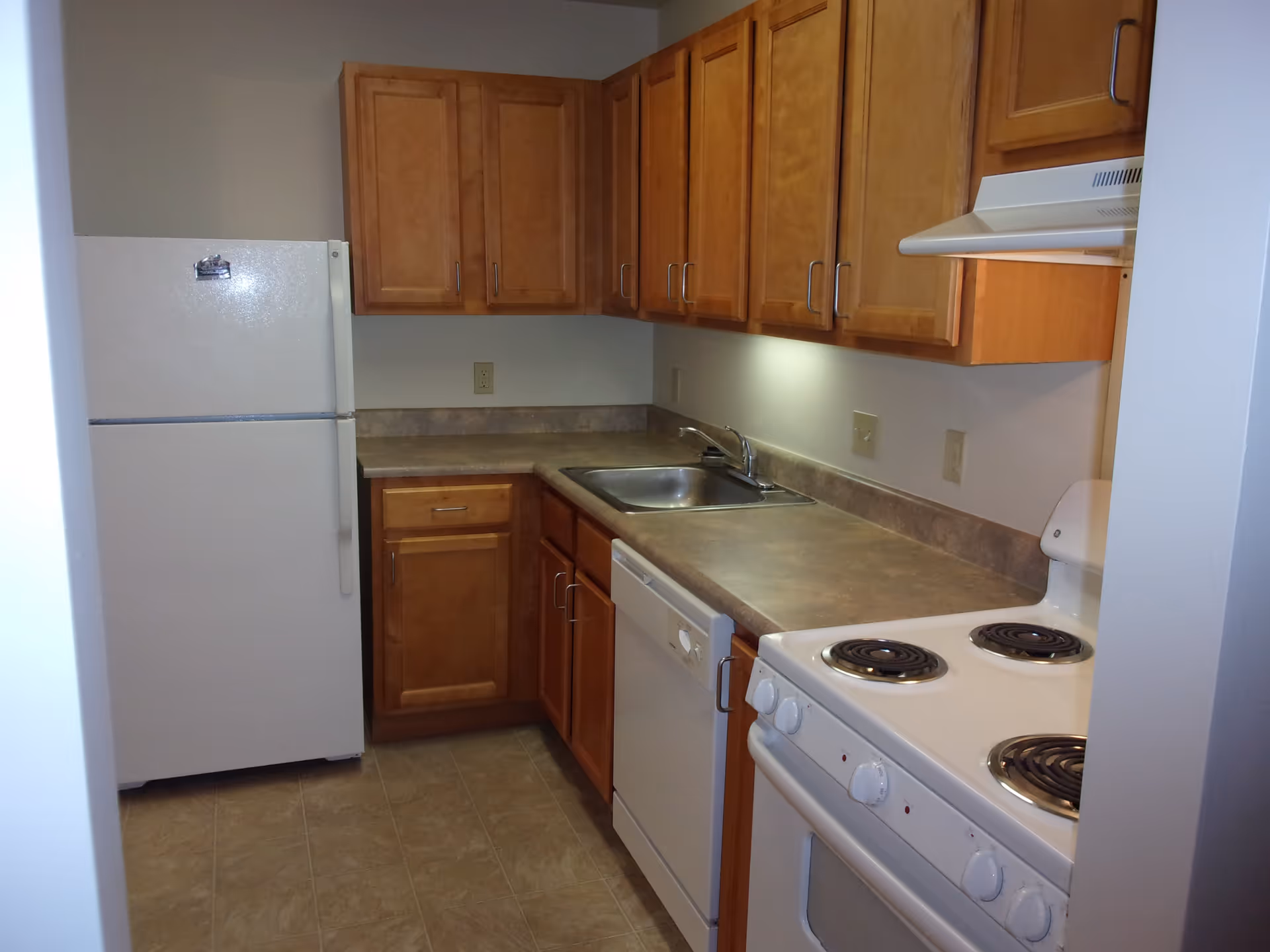 A small kitchen with wooden cabinets, a white refrigerator, a white electric stove with four coil burners, a white dishwasher, a stainless steel sink, and a beige countertop. The floor is tiled and the walls are painted light gray.