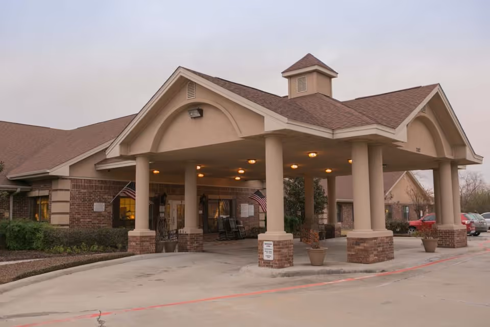 Exterior view of a healthcare and rehabilitation facility entrance with a covered drop-off area supported by columns. The building has a brick and beige facade with a peaked roof and American flags near the entrance. Several cars are parked nearby under an overcast sky.
