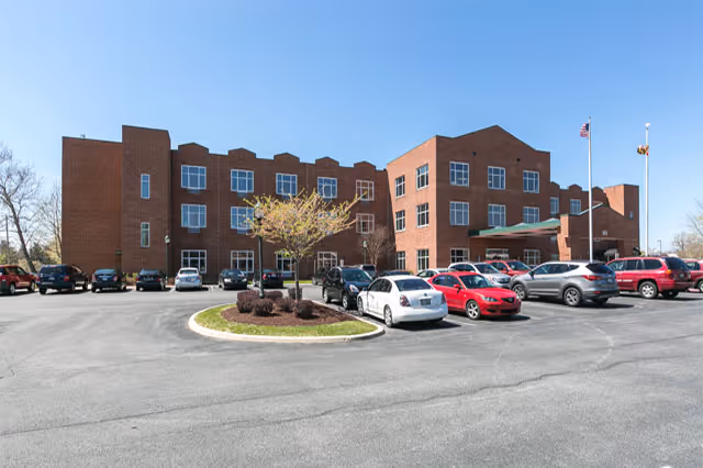 Three-story red brick nursing and rehabilitation building with a front parking lot and cars under a clear blue sky.