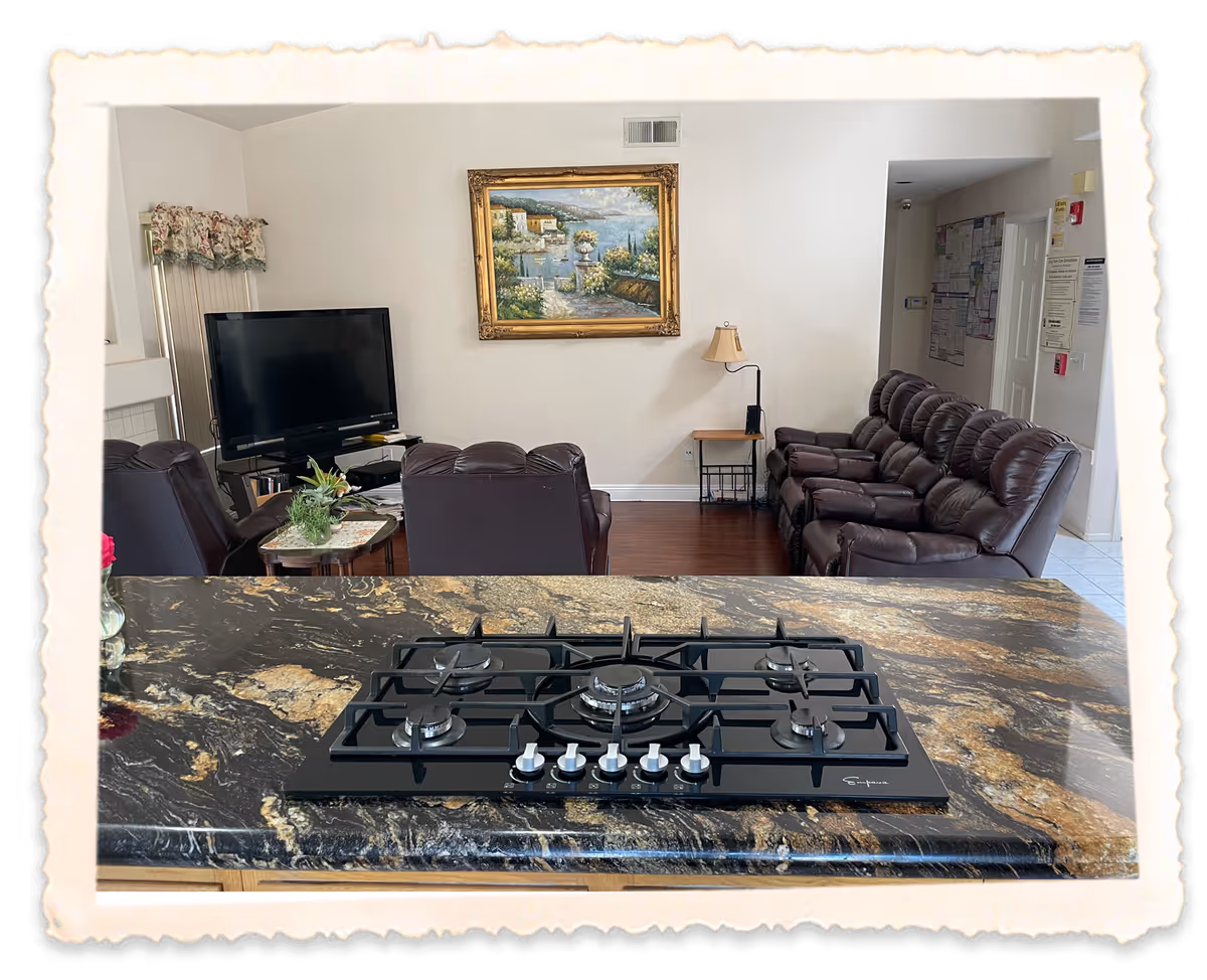 View from a kitchen island with a gas cooktop looking into a living room with leather recliners, a TV, and a framed painting.