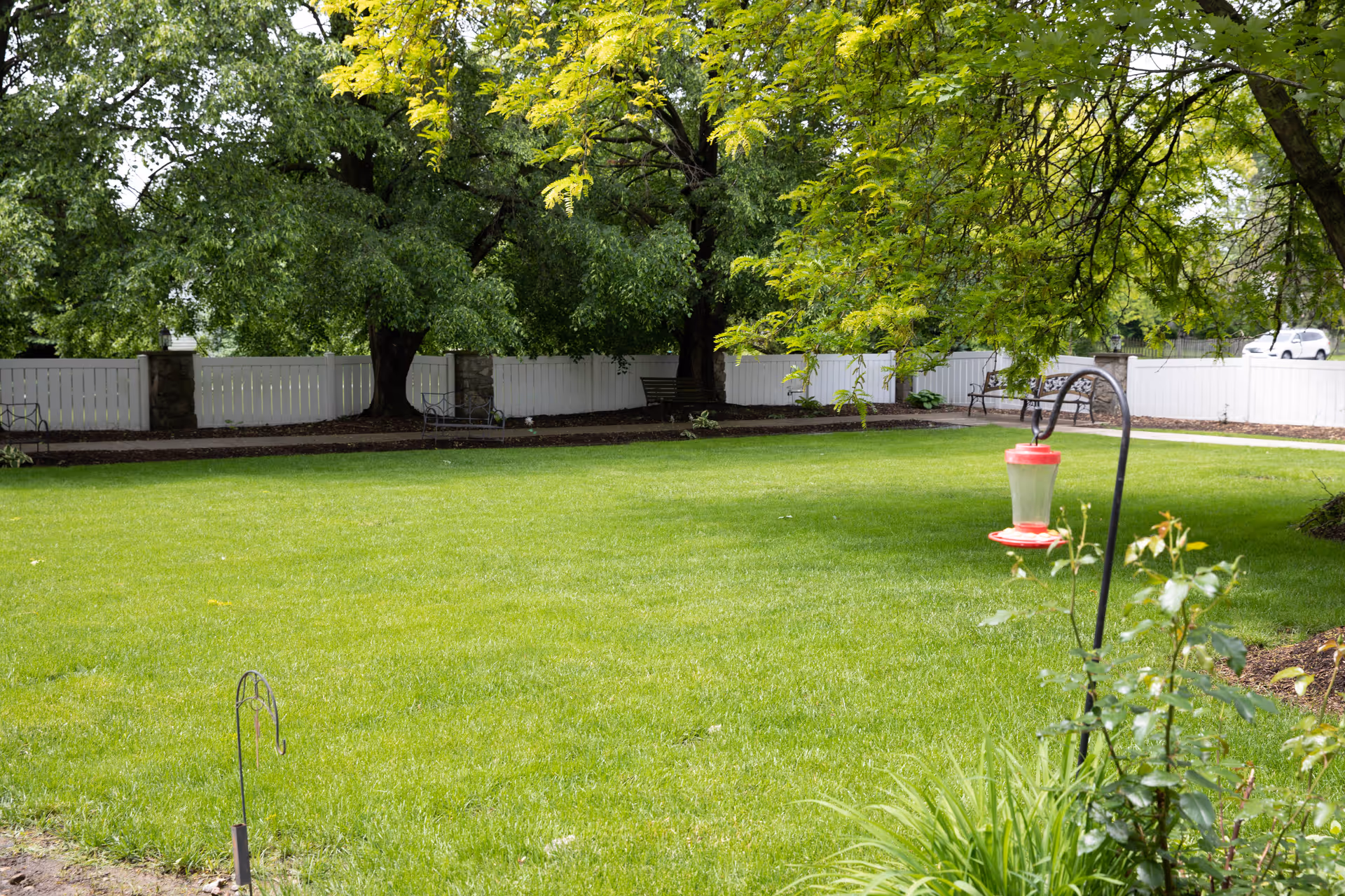 A well-maintained green lawn with a bird feeder hanging from a metal hook in the foreground. The background features large leafy trees, a white wooden fence with stone pillars, and several benches along a paved pathway.