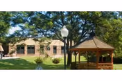 Outdoor area of a senior living facility with a wooden gazebo, green grass, trees providing shade, and a brick building in the background under a clear sky.