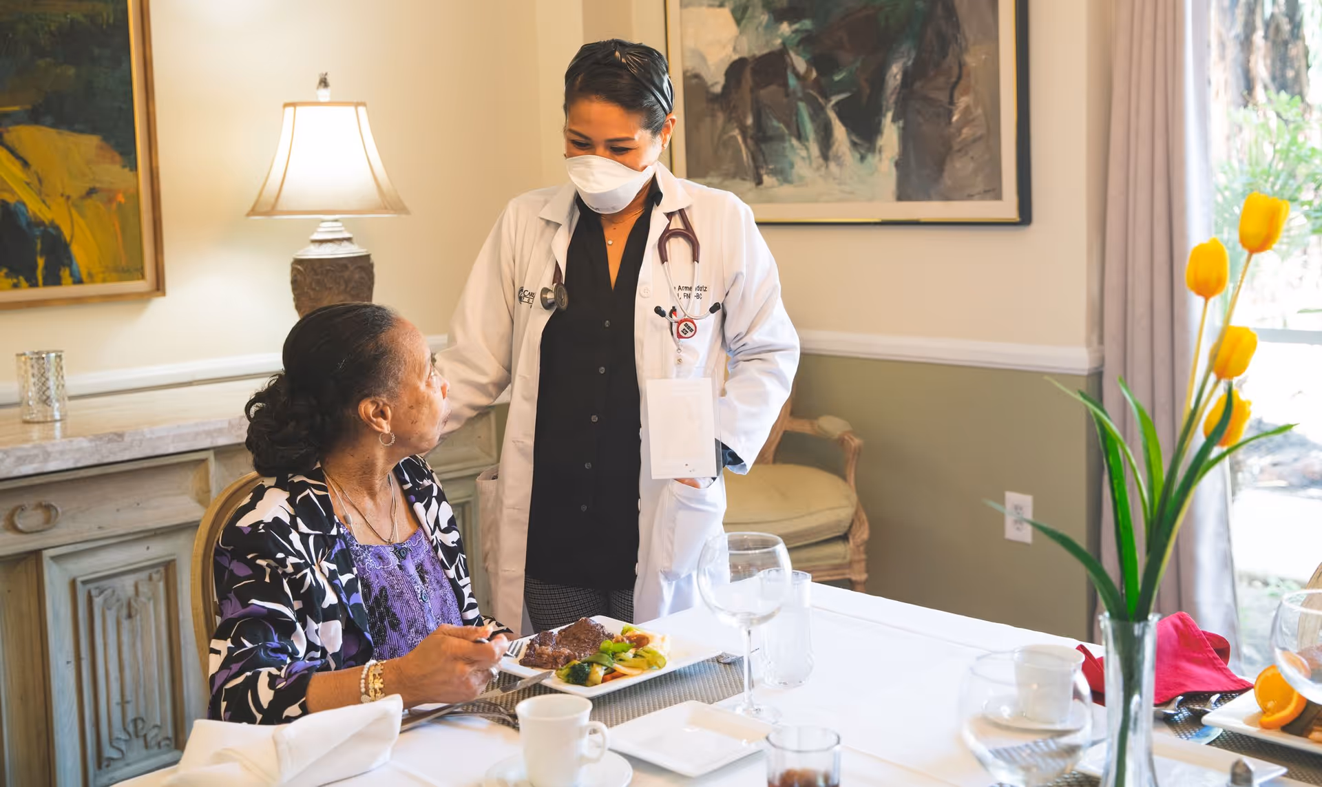 A healthcare professional wearing a white coat and face mask is standing and talking to an elderly woman seated at a dining table with a plate of food. The setting is a well-lit dining room with a lamp, artwork on the wall, and a vase with yellow tulips on the table.