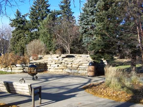 Outdoor patio area with a wooden bench, a charcoal grill, a trash can, and a stone retaining wall surrounded by tall evergreen trees and other vegetation under a clear blue sky.