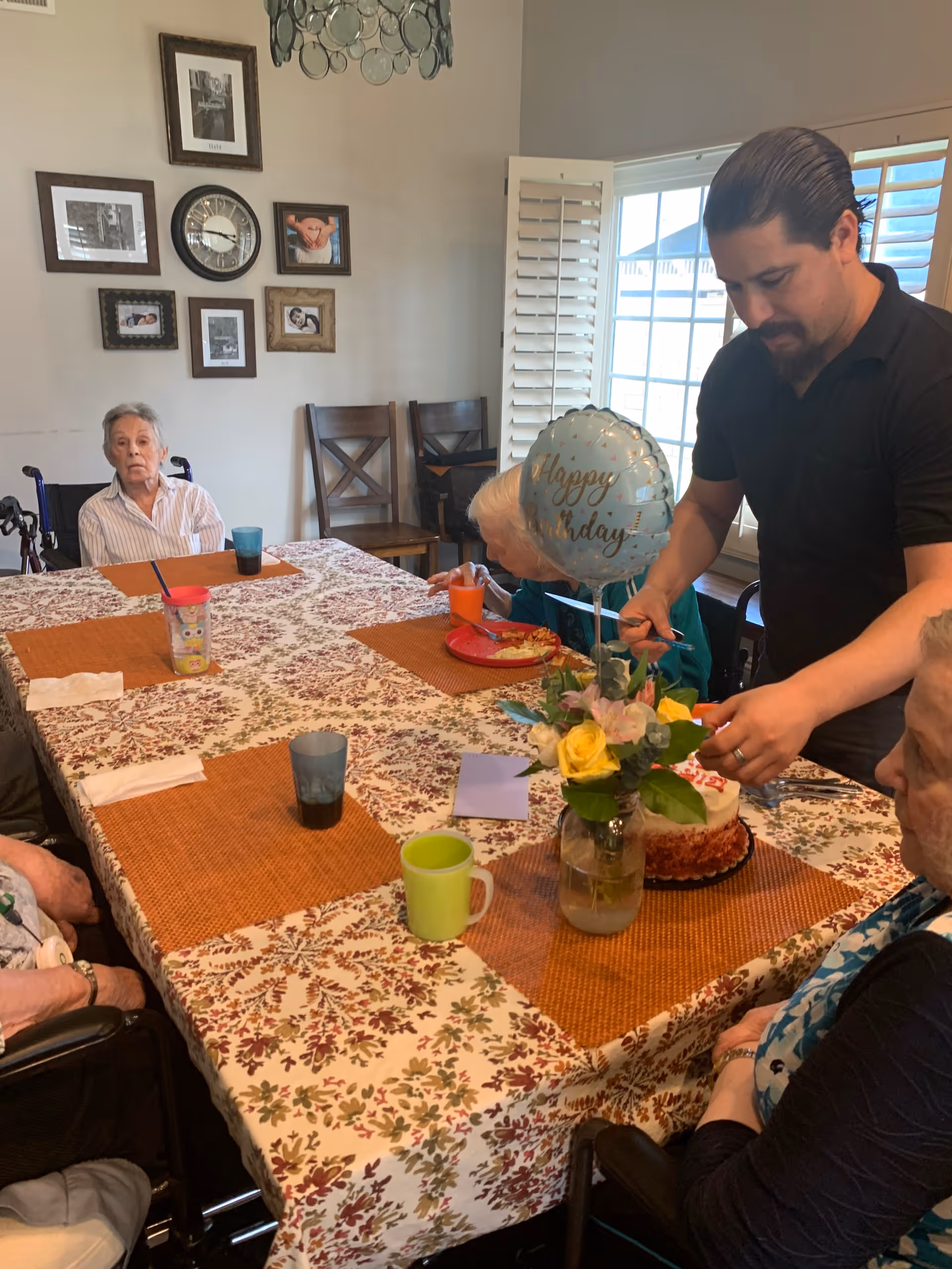 Residents and a staff member gather around a decorated dining table as he cuts a birthday cake under a "Happy Birthday" balloon.