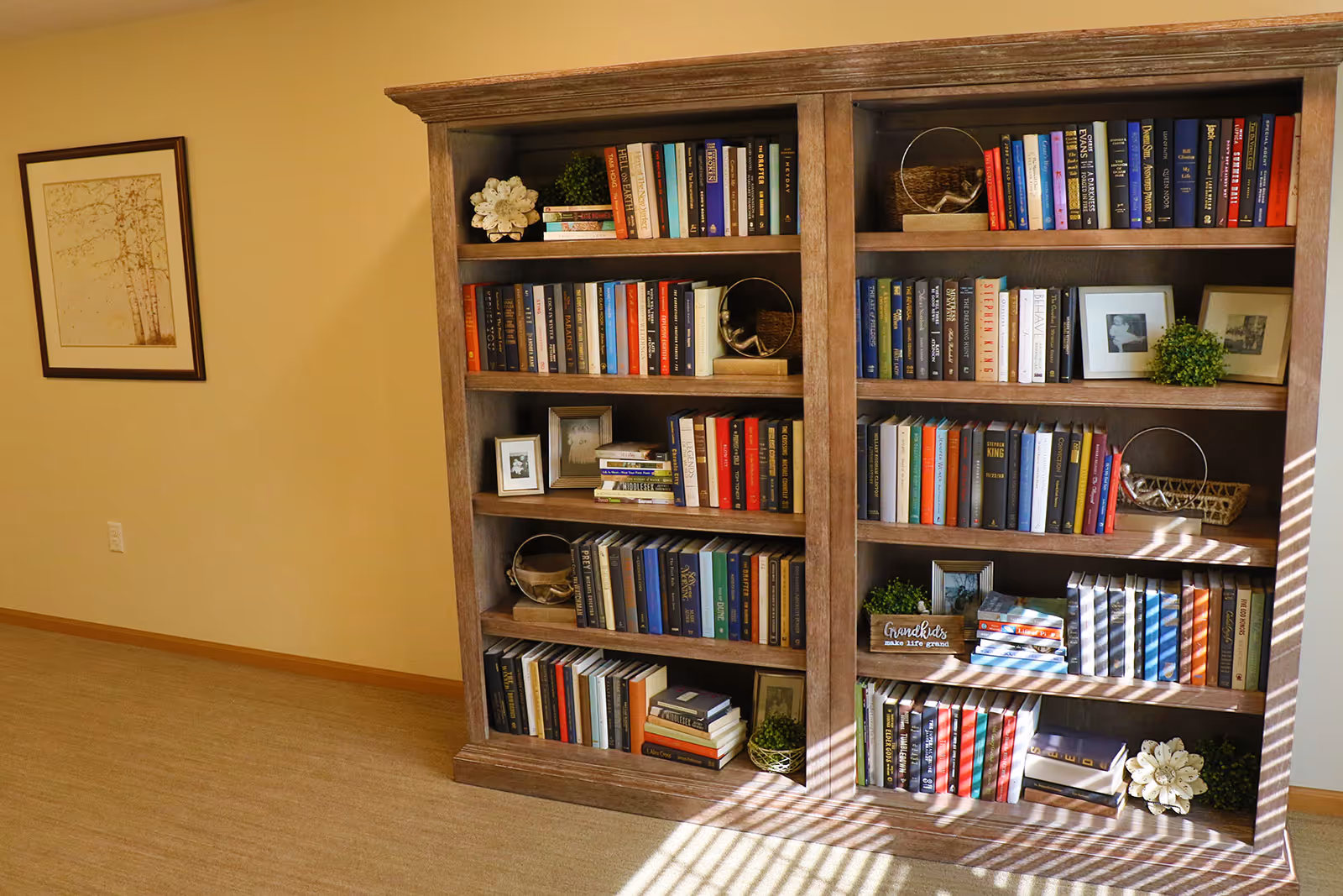 Wooden bookcase filled with books and decorative items against a beige wall with framed art and sunlight on the floor.