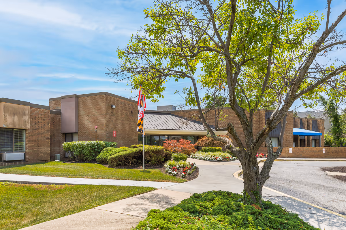 Exterior view of Autumn Lake Healthcare at Patuxent River showing a brick building with a metal roof entrance, landscaped bushes, flowers, a tree in the foreground, and a flagpole with flags.