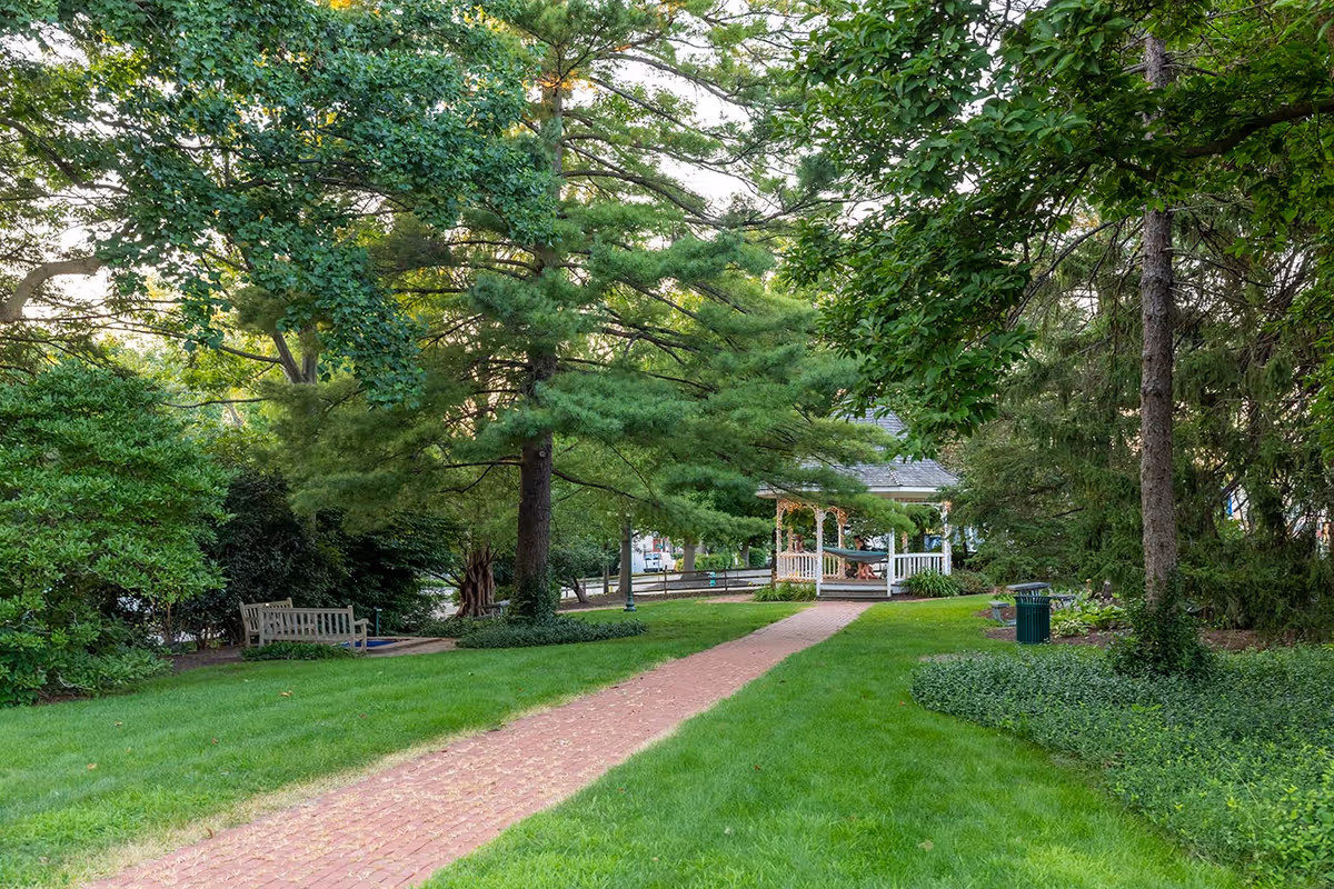 A peaceful outdoor garden area with a red brick pathway leading to a white gazebo surrounded by lush green trees and bushes. There are wooden benches along the path and a green trash can on the right side.