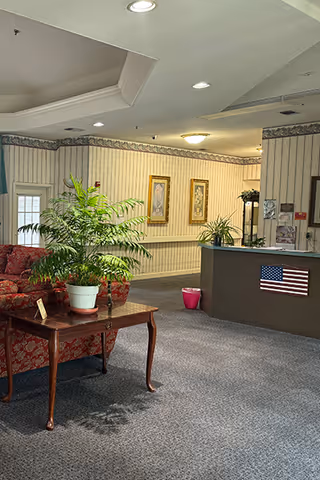 Interior view of a senior living facility lobby area with a red patterned sofa, a wooden coffee table holding a potted plant, a reception desk with an American flag decoration, and framed artwork on the wallpapered walls.