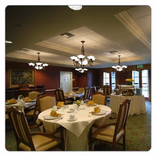 A warmly lit dining room with round tables covered in white tablecloths, set with plates, cups, napkins, and silverware. The room features upholstered wooden chairs, chandeliers hanging from the ceiling, and large windows letting in natural light. A buffet table with covered dishes is visible near the windows.