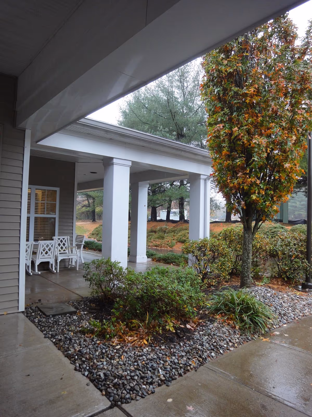 Covered entrance with white columns, a porch with chairs, and landscaped shrubs and a tree beside a wet walkway.