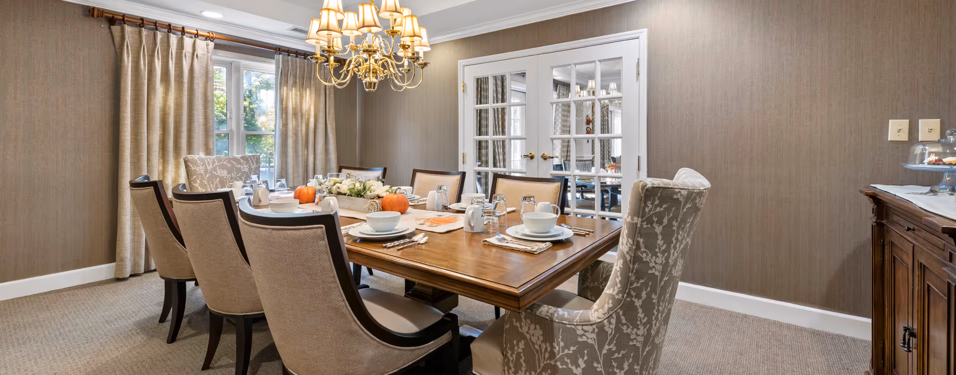 A well-lit dining room with a wooden dining table set for eight people. The table is decorated with white plates, bowls, cups, silverware, glassware, a floral centerpiece, and small pumpkins. The room features beige walls, a beige carpet, beige curtains covering a window, a chandelier with multiple lampshades hanging above the table, and white French doors with glass panes. A wooden sideboard with a glass cake stand is visible on the right side.