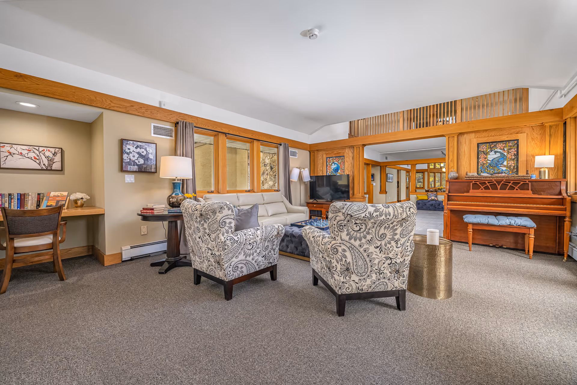 A cozy living room area with two patterned armchairs facing a beige sofa and a TV on a wooden stand. The room features wooden trim and a piano with a cushioned bench. There is a small desk with a chair and books on the left side, and decorative stained glass panels above the piano. The carpeted floor and neutral walls create a warm and inviting atmosphere.