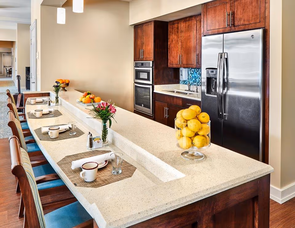 A modern kitchen area with a long beige countertop featuring place settings with cups, glasses, and napkins on woven placemats. There are wooden chairs with blue cushions along the counter. The kitchen has dark wooden cabinets, a stainless steel refrigerator, built-in oven and microwave, and a small sink. Decorative elements include a glass bowl filled with lemons and a vase with pink flowers on the counter.