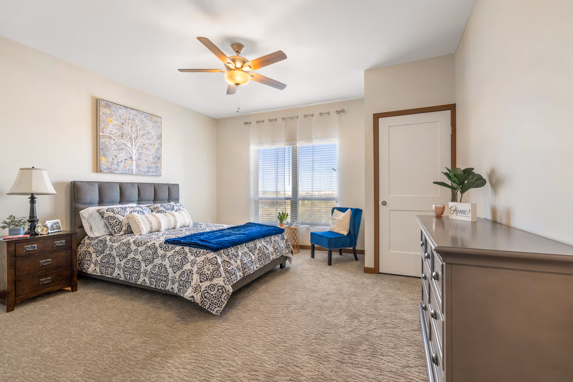 Bright, furnished bedroom with a bed, nightstand, dresser, blue accent chair, and ceiling fan.