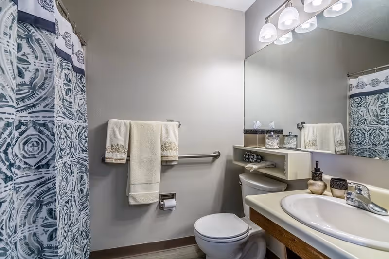A clean and well-lit bathroom featuring a toilet, a sink with a countertop, a large mirror above the sink, and a shower with a patterned blue and white curtain. There are two cream-colored towels hanging on a metal towel rack above the toilet paper holder, and decorative items on the sink countertop and a small shelf above the toilet.