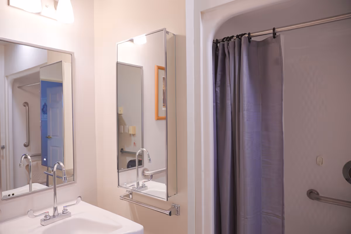 A bathroom with a white sink and chrome faucet, two rectangular mirrors above the sink, a gray shower curtain on a curved shower rod, and grab bars inside the shower area.
