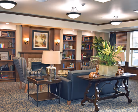Cozy communal lounge with sofas and armchairs facing a fireplace, flanked by bookshelves and a decorative table with a large plant.