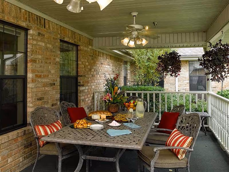 Covered outdoor patio area with a metal table set for snacks including croissants, cookies, cheese, and lemonade. Four cushioned chairs with red and striped pillows surround the table. Hanging plants and a ceiling fan with lights are visible, along with brick walls and windows of the building.