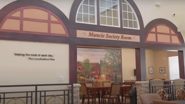 Interior view of the Muncie Society Room at Lincolnshire Place of Decatur, featuring a large mural of a rural farm scene with a barn and silo. The room has wooden framed arches with windows above, a round table with chairs, and a quote on the wall that reads 'Making the most of each day... The Lincolnshire Way.'