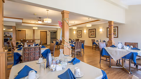 A spacious dining room with multiple tables covered in beige tablecloths and blue napkins. Each table is set with cups, glasses, and condiments. The room features wooden floors, light-colored walls, decorative columns wrapped with garlands, and framed artwork on the walls. Ceiling lights provide warm illumination.