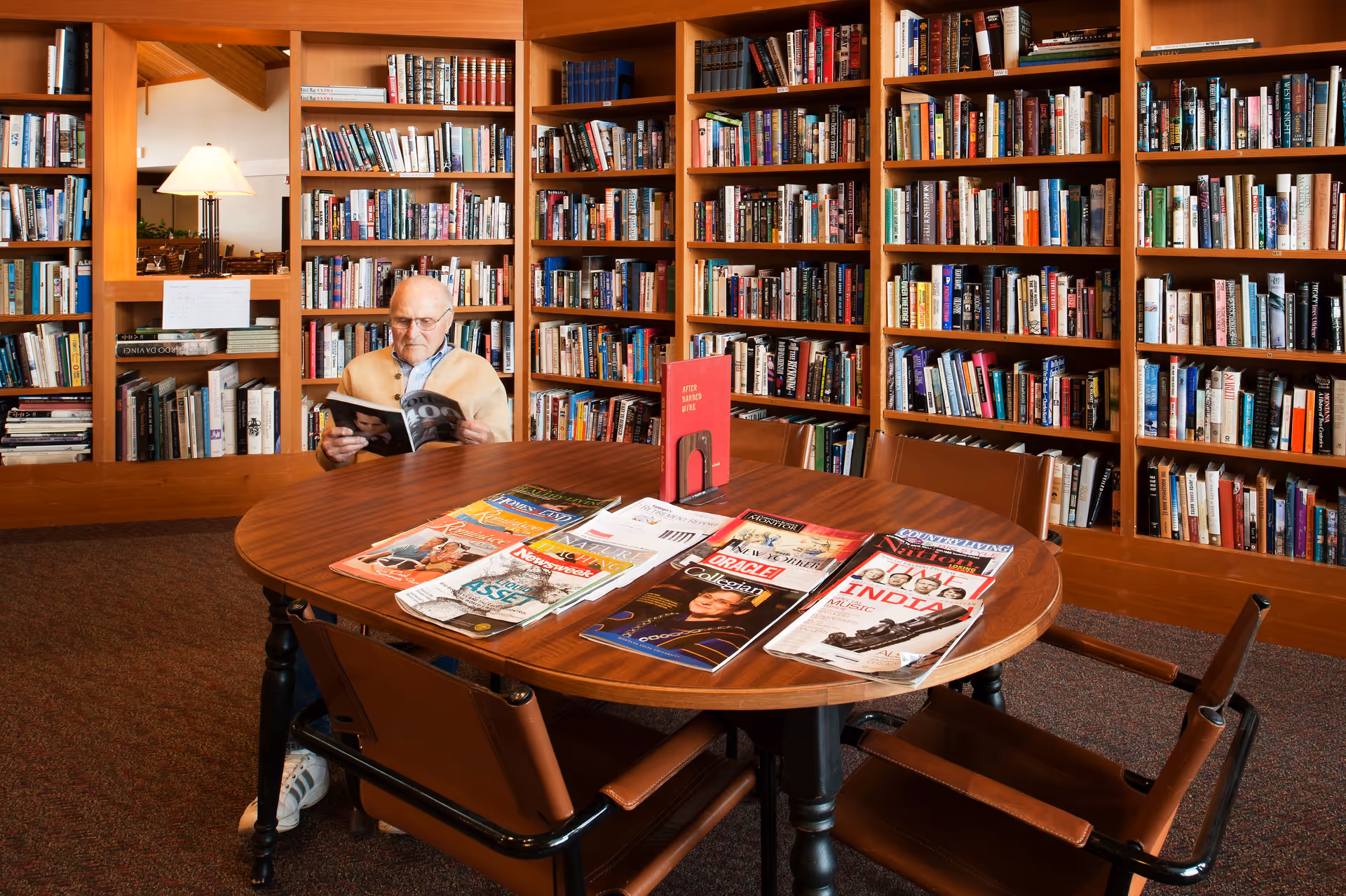 An elderly man sitting at a round wooden table in a library room, reading a magazine. The table has several magazines spread out on it. Behind him are tall wooden bookshelves filled with books. The room has warm lighting and a cozy atmosphere.