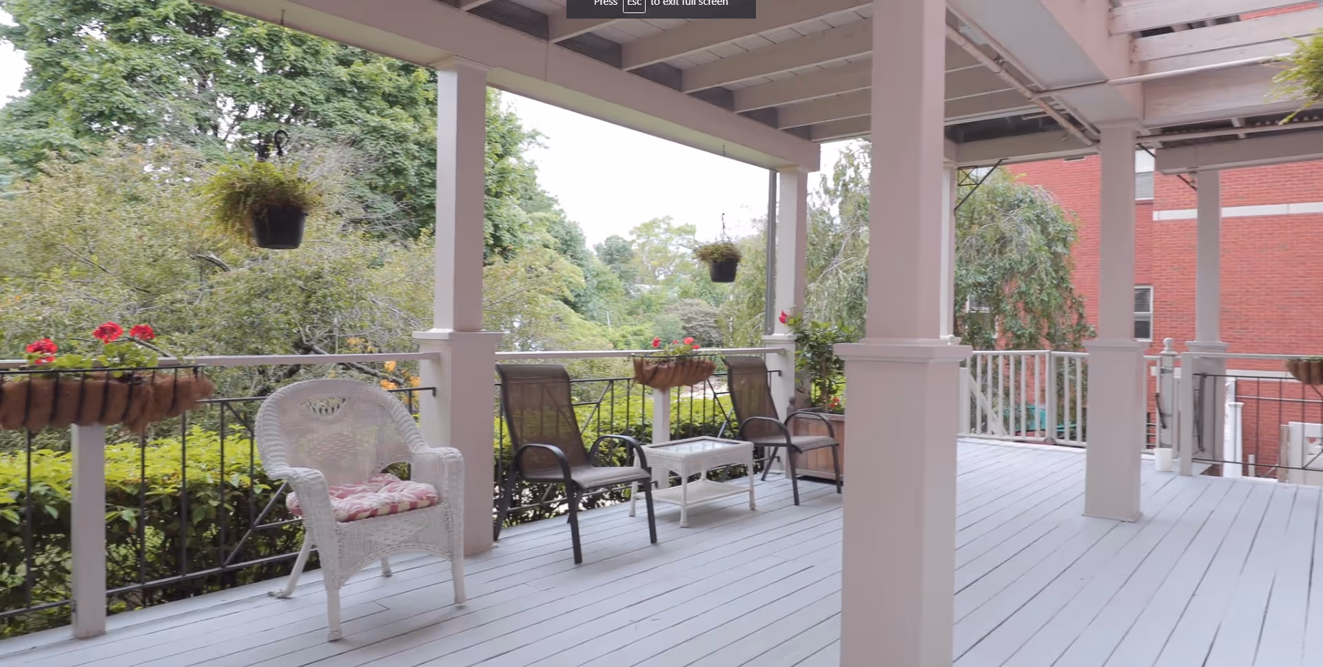 Covered outdoor porch area with white wooden floor and columns, featuring a white wicker chair with a cushion, two brown chairs, a small white table, hanging flower pots, and railing with flower boxes. Green trees and a red brick building are visible in the background.