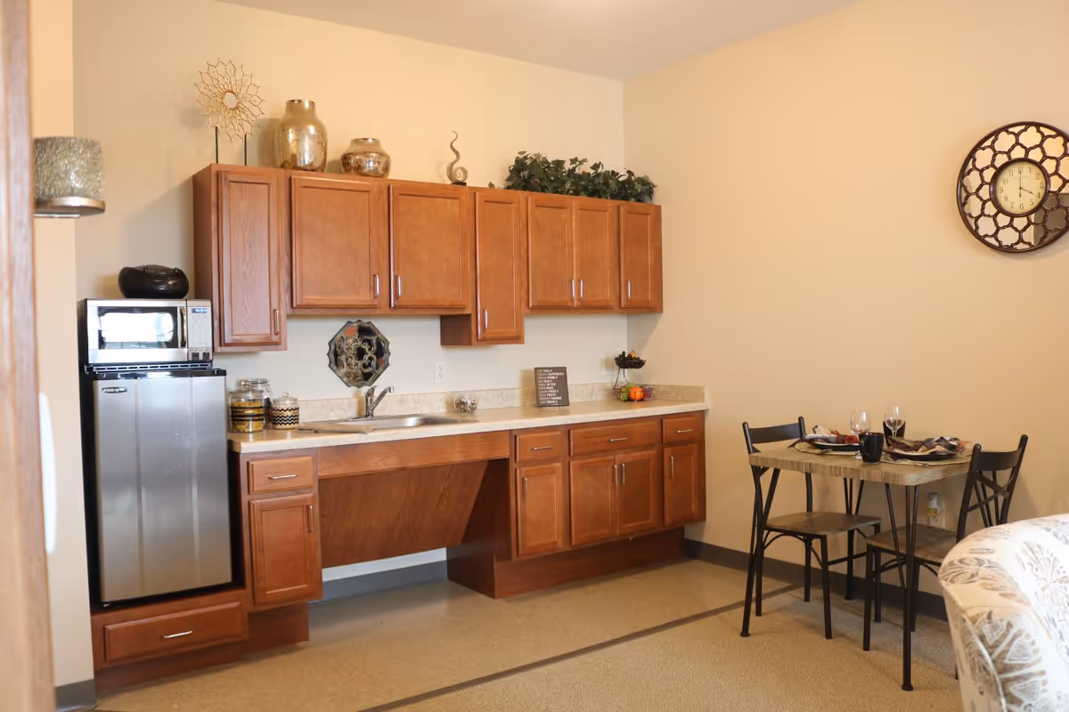 A small kitchen area in a senior living facility with wooden cabinets, a countertop with a sink, a mini refrigerator, and a microwave. There are decorative items on top of the cabinets and a small dining table with two chairs set with plates, glasses, and napkins. A decorative wall clock is mounted on the beige wall.