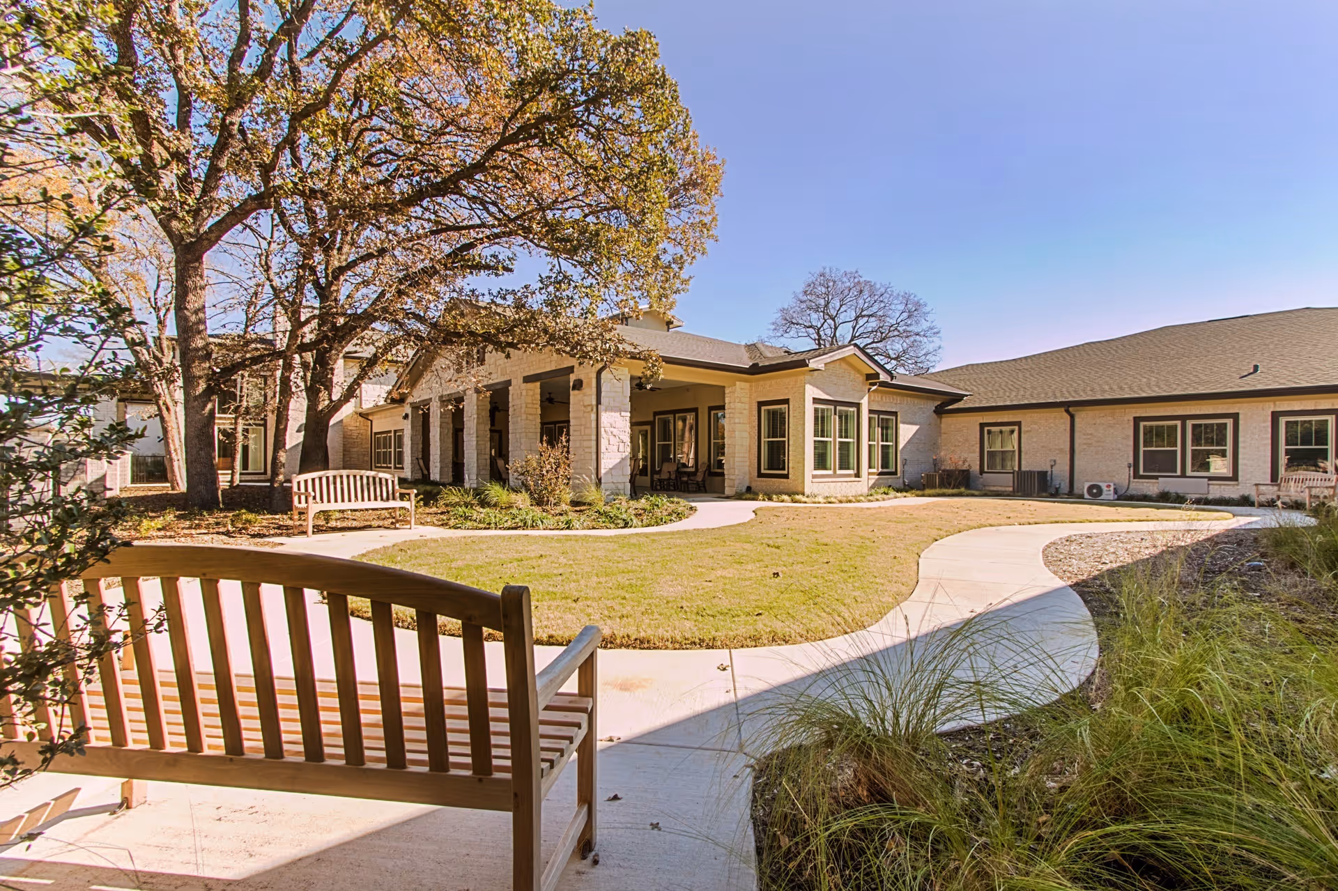 Outdoor courtyard area at The Oaks at Flower Mound featuring wooden benches, a curved concrete walkway, grass, trees, and a single-story building with multiple windows under a clear blue sky.